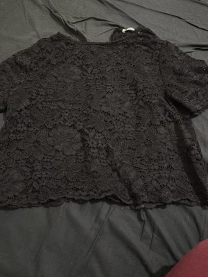 Svart blommig spetsblus med dragkedja - Säljer en svart blus i genombruten spets med blommigt mönster. Toppen har korta ärmar och dragkedja i ryggen. Perfekt för dig som gillar detaljer och vill ha en snygg och stilren look. Materialet är mjukt och följsamt.Det är storlek s men passar m också den är väldigt snygg men inte min stil längre och har bara använt den 1 gg. Köpte den också utomlands så vet inte vad det är för varormärke om jag ska vara ärlig 
