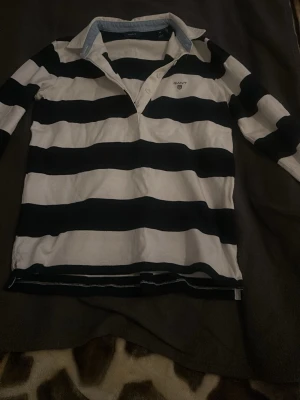 Randig långärmad henley från GANT - Säljer en klassisk långärmad henley från GANT i storlek XS. Skjortan har breda svarta och vita ränder, krage med ljusblå insida och knappar framtill. GANT-logga med broderad sköld på bröstet. Helt ny 