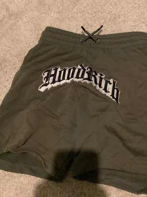 Olivgröna Hoodrich shorts med snörning - Säljer ett par olivgröna shorts från Hoodrich med stor broderad logga framtill och textdetalj bak. Shortsen har elastisk midja med snörning och en bakficka. Perfekta för chill dagar eller streetwear-stil.
