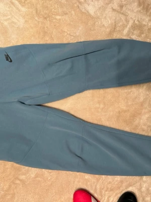 Nike tech byxor M - Säljer ett par blå mjukisbyxor från Nike i storlek M. Byxorna har en enkel design med svart Nike-logga på låret och elastisk midja med dragsko. Perfekta för chill eller träning, tillverkade i mjukt material för extra komfort.