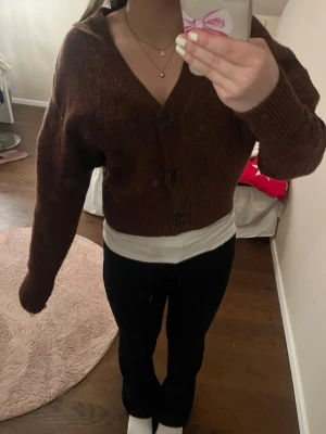 Brun croppad kofta med knappar - Mysig brun croppad kofta med tre stora knappar framtill och v-ringad hals. Koftan har långärmad passform och är stickad i ett mjukt material och sticks inte. Perfekt att slänga över en topp för en chill look.