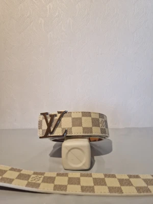 Rutig beige LV-bälte - Säljer ett beige och brunt rutigt bälte från Louis Vuitton med den klassiska LV-loggan i silver som spänne. Bältet har ett stilrent och lyxigt uttryck, perfekt för att lyfta vilken outfit som helst.