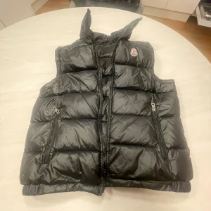  Moncler väst - Svart dunväst från Moncler med quiltad design och hög krage. Västen har två dragkedjefickor framtill och stängs med dragkedja. Klassisk Moncler-logga på bröstet. liten spricka där bak men syns knappt, Perfekt för lager-på-lager och snygg till streetwear.