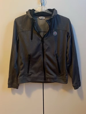 Grå kofta Moncler  - Säljer en grå kofta från Moncler i storlek S. Jackan har huva med snörning, två fickor med dragkedja och klassisk Moncler-logga på bröstet. Perfekt för dig som vill ha en stilren och sportig look.