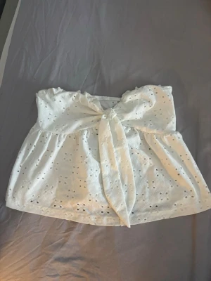 Vit axelbandslös broderad tubtopp - Säljer en söt vit axelbandslös tubtopp med broderade hålmönster och stor knytrosett framtill💞