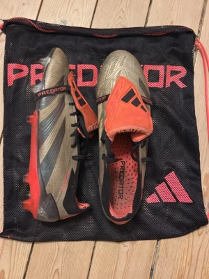 Adidas Predator fotbollsskor grå/röd/carbon - Säljer ett par Adidas Predator fotbollsskor i grå, svart och röd färg. Skorna har snörning, täckt plös och mönstrad ovandel för extra grepp. Sulan har fasta dobbar och tydlig Predator-logga. Perfekta för dig som vill sticka ut på planen. Orginal boxen och sko påse kommer även med!