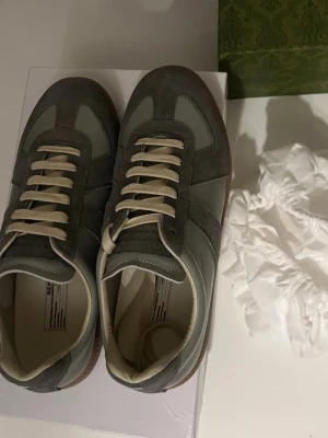 Gråa Maison Margiela  - Säljer ett par stilrena sneakers från Maison Margiela i grå mocka och skinn med vita snören och gummisula. Klassisk siluett med rund tå och passar perfekt med allt. Fick dem i present. Pris kan diskuteras det är bara och skriva vid intresse. Kvitto finns med🍾