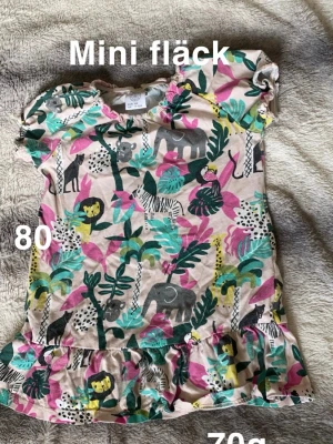 Färgglad djurmönstrad kortklänning - Supersöt kortklänning från Lindex med färgglatt djurmönster i rosa, turkos, gul och svart. Klänningen har volang nertill och korta ärmar. Perfekt för dig som gillar lekfulla prints. Mini fläck. Storlek 80