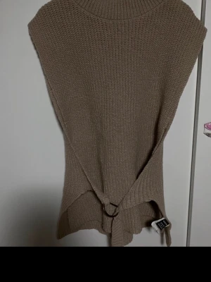 Beige stickad väst med knyt - Säljer en beige stickad väst med hög krage. Västen har slitsar i sidorna och ett snyggt knytband med metallring i midjan för extra detalj. Perfekt att slänga över en skjorta eller t-shirt för en chill look.