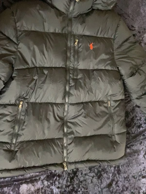Mörkgrön pufferjacka från Polo Ralph Lauren - Säljer en mörkgrön pufferjacka från Polo Ralph Lauren med ikonisk röd logga på bröstet. Jackan har flera dragkedjeförsedda fickor, hög krage och quiltad design. Perfekt för kalla dagar och riktigt snygg till streetwear-stilen.