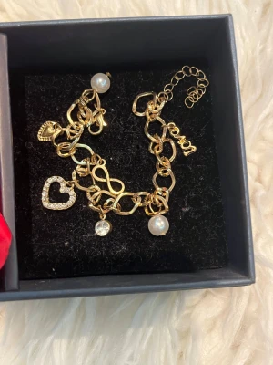Guldigt armband med pärlor och berlocker - Säljer ett snyggt armband i guldfärgad metall med flera berlocker, bland annat pärlor, ett hjärta med stenar och små detaljer som löv och text. Perfekt accessoar för att lyfta din outfit.