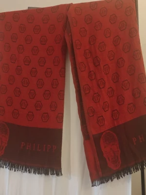 Röd Philipp Plein halsduk med döskalle - Snygg röd halsduk från Philipp Plein med all-over logomönster och stora döskallar i ändarna. Halsduken har fransar i kanterna och är tillverkad i ett mjukt material som känns lyxigt. Perfekt accessoar för att lyfta din outfit. Nypris 4500- 5000