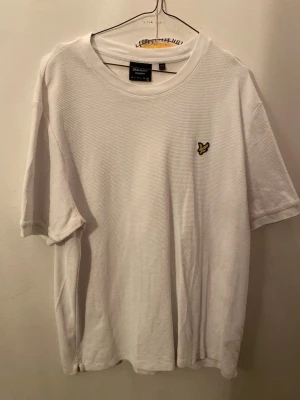 Vit relaxed fit t-shirt Lyle & Scott - Vit t-shirt från Lyle & Scott i relaxed fit med klassisk rund hals och korta ärmar. Materialet är bomull med en diskret struktur och bröstet pryds av den ikoniska gula fågel-loggan. Enkel och clean stil som funkar till allt.