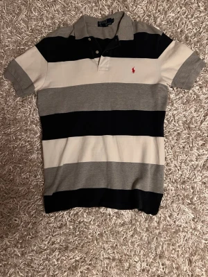 Randig pikétröja från Polo Ralph Lauren - Säljer en klassisk pikétröja från Polo Ralph Lauren i storlek S. Tröjan har breda ränder i svart, grått och vitt samt en röd broderad logga på bröstet. Kortärmad med krage och knappar framtill. Perfekt för en stilren och avslappnad look.