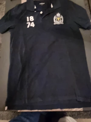 Mörkblå pikétröja Lyle & Scott M - Snygg mörkblå pikétröja från Lyle & Scott med broderad logga och siffrorna 1874 på bröstet. Klassisk krage med två knappar och vita detaljer. Perfekt till en avslappnad stil och passar till jeans eller shorts.