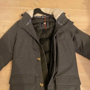 Grå parkas från Seaside XS - Mörkgrå parkas från Seaside med mysig vit teddyfoder i huvan. Jackan har stora fickor framtill, dragkedja och knappar. Perfekt för kalla dagar och har en klassisk, rak passform. Snygg och enkel design som funkar till det mesta.