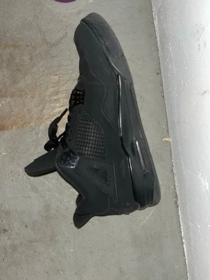 Svarta Nike Air Jordan 4 sneakers - Säljer ett par helsvarta Nike Air Jordan 4 sneakers med klassisk meshpanel på sidan, Jumpman-logga på häl och sula samt snygga detaljer i syntet och mocka. Skorna har snörning och en chunky sula som ger grym streetstil.