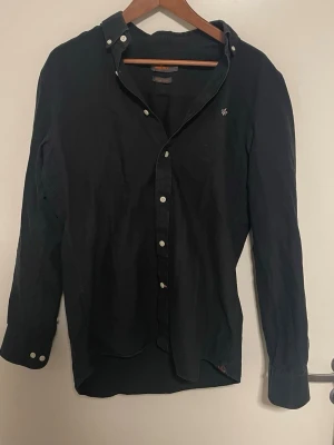 Svart skjorta från Morris Stockholm - Klassisk svart skjorta från Morris Stockholm i storlek M, tillverkad i bomull med diskret broderad logga på bröstet. Skjortan har knappar hela vägen och button down-krage. Perfekt till jeans eller chinos för en clean look.