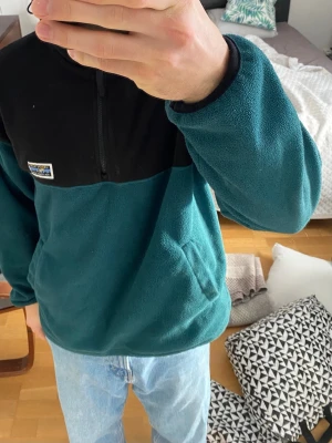 Svart och grön fleecehoodie Junkyard M - Mysig hoodie från Junkyard i svart och mörkgrön fleece. Tröjan har huva, halv dragkedja framtill och en liten patch på bröstet. 