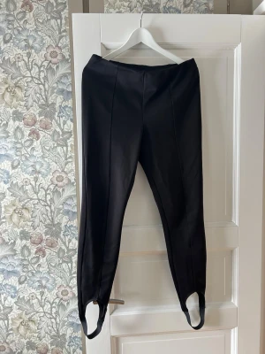 Svarta stretchiga byxor med fotstrap M - Säljer ett par svarta byxor i storlek M med smal passform och praktiska fotstrappar. Byxorna är sydda i ett stretchigt material av 96% polyester och 4% elastan, vilket gör dem riktigt bekväma. Snygga sömmar framtill ger en clean look.