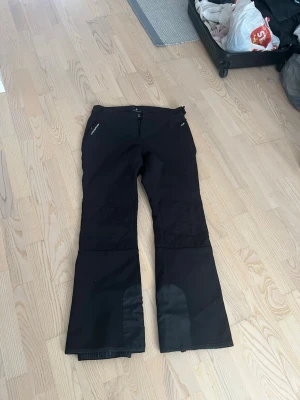 Svart täckbyxa, långa ben och bred högre midja - Säljer ett par svarta täckbyxor, softshell från Cross Sportswear i storlek L. Byxorna har sidofickor med dragkedjor, förstärkta partier vid bensluten och fleecefoder på insidan för extra värme. Perfekta för vintern och outdoor-aktiviteter.med dragkedja bak ben för att täcka pjäxor i backen. Mjt sköna på. 