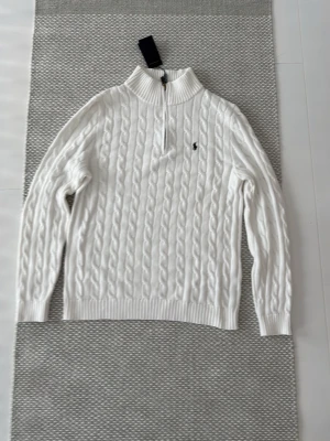 Vit kabelstickad tröja Polo Ralph Lauren - Säljer en vit kabelstickad tröja från Polo Ralph Lauren i storlek M. Tröjan har half zip, hög krage och den klassiska broderade loggan på bröstet. Tillverk bi bomull och har ribbade muddar vid ärmar och nederkant w. Perfekt för en clean och stilren look.