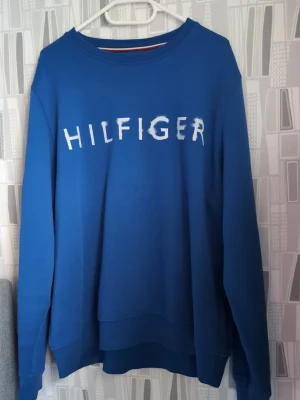 Blå sweatshirt från Tommy Hilfiger XXL - Säljer en blå sweatshirt från Tommy Hilfiger i storlek XXL. Tröjan har ett stort vitt Hilfiger-tryck på bröstet och klassisk rund hals. Materialet är mjukt och skönt bomull, med långa ärmar och ribbade muddar. Perfekt för chill dagar och enkel stil.