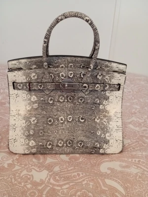 B30 exotic lizard  - Säljer en lyxig handväska i svartvitt mönstrat skinn med metallspänne framtill. Väskan har två rejäla handtag och ett unikt, nästan ormliknande mönster. Insidan är ljusbeige och väskan är rymlig med klassisk form. Perfekt för dig som vill sticka ut med en statementväska.