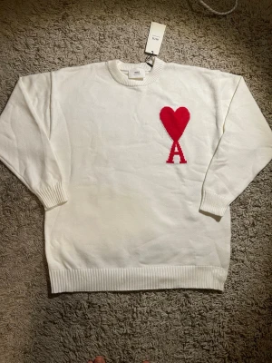 Vit stickad tröja från Ami med rött hjärta - Vit stickad tröja från Ami med rund halsringning och ribbade muddar. På bröstet finns ett stort rött hjärta och bokstaven A. Tröjan har en clean look och är perfekt för dig som gillar stilrena detaljer.