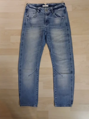 Blå jeans från Lindex, storlek 36 - Snygga blå jeans från Lindex i storlek 36 med klassisk femficksdesign och ljusa slitningar framtill och baktill. Jeansen har rak passform och normal midja, tillverkade i bomullsmaterial för en skön känsla. Perfekt för dig som gillar enkel och stilren denim.
