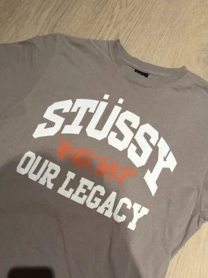 Stüssy x Our Legacy grå t-shirt - Snygg grå t-shirt från Stüssy x Our Legacy Workshop med stor vit och orange print på bröstet. Klassisk rund hals och korta ärmar. Tillverkad i mjuk bomull som är skön mot huden. Perfekt för dig som gillar streetwear och vill sticka ut med en unik collab.