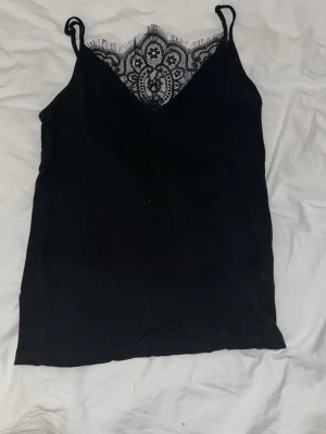Svart linnetopp med spetsdetaljer - Säljer en svart linnetopp med tunna axelband och snygg spets upptill i ett dekorativt mönster. Toppen har v-ringad hals och ärmlös design, perfekt för att styla med jeans eller kjol. Materialet känns mjukt och lätt mot huden.