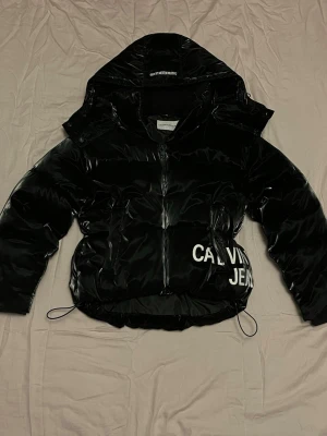  Calvin Klein pufferjacka  - Säljer en svart, glansig pufferjacka från Calvin Klein med stor avtagbar huva och vit logga på sidan. Jackan är köpt second hand för cirka två år sedan men är i bra skick utan defekter. Storlek S. Ge gärna prisförslag!😇