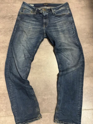 Blå jeans från Jack & Jones, storlek 34/32 - Säljer ett par klassiska blå jeans från Jack & Jones Jeans Intelligence. De har fem fickor, normal passform och snygga slitningar framtill för en avslappnad look. Jeansen är i denim med kontrastsömmar och har en skinnpatch bak i midjan.