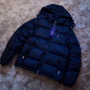 Svart pufferjacka från Polo Ralph Lauren - Säljer en svart pufferjacka från Polo Ralph Lauren med huva och två dragkedjeförsedda fickor. Jackan har det klassiska röda Polo-logot på bröstet och är vadderad för extra värme. Perfekt för kalla dagar och har en clean, stilren look. QR-kod funkar. Ställ frågor vid fundering 😊