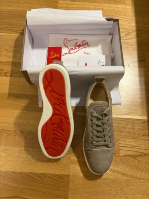 Beige sneakers från Christian Louboutin - Snygga beige sneakers från Christian Louboutin i mocka med klassisk röd sula och vita kanter. Skorna har rund tå, snörning och en stilren design som passar till det mesta. Perfekta för dig som vill ha något exklusivt och trendigt.