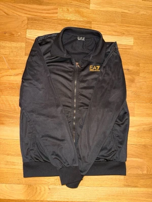 Svart bomberjacka från EA7 Emporio Armani - Säljer en svart bomberjacka från EA7 Emporio Armani med gul logga på bröstet. Jackan har dragkedja framtill, ribbade muddar vid ärmslut och midja samt två sidofickor. Perfekt för dig som vill ha en stilren och sportig look. 