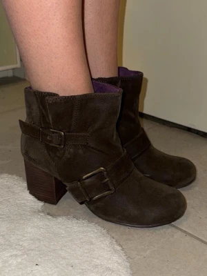 Bruna mockaboots med spännen - Snygga bruna boots från Blowfish i mjuk mocka med grov klack och dekorativa metallspännen. Insidan är fodrad i lila tyg för en cool kontrast. Rund tå och dragkedja på insidan gör dem enkla att ta på. Perfekta till höstens outfits!