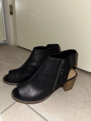 Svarta stövletter med öppen tå och klack - Snygga svarta stövletter i skinnimitation med öppen tå och låg klack i trälook. Skorna har dragkedja på sidan och en stilren design som passar till många outfits. Perfekta för dig som vill ha något både trendigt och bekvämt. Klackarna är helt nya‼️