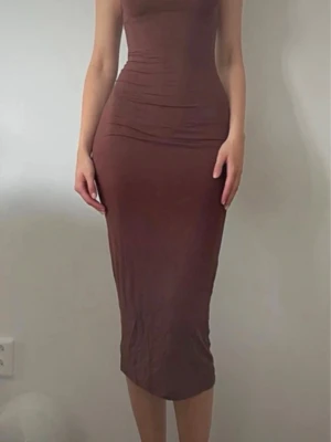 Brun långklänning med öppen rygg - Säljer en brun långklänning med smala axelband som går att  korsas i ryggen men som även går att bära vanligt. Klänningen är figurnära och har en enkel, stil