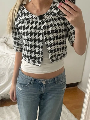 Svartvit rutig kofta/bolero från Jsfn - Säljer en croppad svartvit kofta från Jsfn med stort rutmönster och en dekorativ knapp framtill. Koftan har trekvartsärm och rund halsringning. Perfekt för dig som vill ha en statement piece till garderoben.