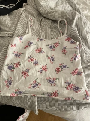 Vit blommig linnetopp med smala band - Supersöt vit linnetopp med tunna axelband och blommigt mönster i lila och rött. Toppen har en rak passform och är tillverkad i ett mjukt, lätt material som passar perfekt till varma dagar. Perfekt att styla med jeans eller kjol för en fräsch look. Den är i ungefär storlek small/medium. 
