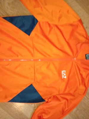 Orange vindjacka från EA7 Emporio Armani och tillhörande byxor - Snygg orange vindjacka från EA7 Emporio Armani med marinblå detaljer. Jackan har dragkedja framtill, lång ärm och stort logotryck på ryggen. Tillverkad i lätt polyester som passar perfekt för en sportig look. Tillhörande byxor till denna tröja!
