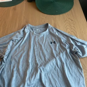 Grå Under Armour Tech Tee t-shirt - Säljer en grå Under Armour Tech Tee t-shirt i storlek L. T-shirten har korta ärmar, rund hals och en svart Under Armour-logga på bröstet. Tillverkad i polyester för en lätt och skön känsla, perfekt för träning eller chill dagar.