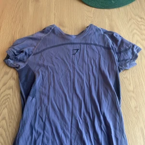 Lila Gymshark t-shirt i storlek S - Snygg lila t-shirt från Gymshark med korta ärmar och diskret logga på bröstet. Tillverkad i en blandning av nylon och polyester som ger en stretchig och ventilerande känsla, perfekt för träning. T-shirten har ribbade detaljer och mönstrad mesh på ryggen för extra komfort.