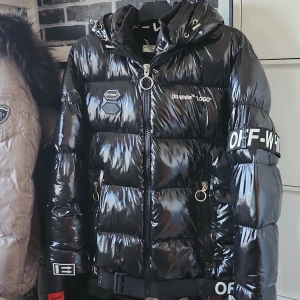 Svart pufferjacka från Off-White - Säljer en svart, glansig pufferjacka från Off-White med huva och feta logodetaljer på bröstet, ryggen och ärmen. Jackan har dragkedja med ring, snörning vid huvan och flera coola Off-White-prints. Perfekt för dig som vill sticka ut i vinter.