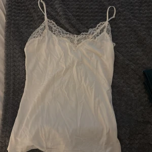 Vit spetskantad linnetopp från Lindex - Supersöt vit linnetopp från Lindex i storlek S. Toppen har tunna axelband och en vacker spetskant med blommigt mönster längs halsringningen. Materialet är mjukt och har en lätt glans, perfekt för att ge en fräsch och feminin vibe.