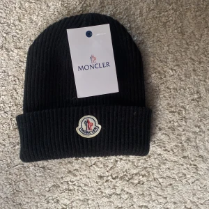 Svart ribbstickad mössa från Moncler - Svart ribbstickad mössa från Moncler med klassisk uppvikt kant och broderad logotyp framtill. Mössan är tillverkad i mjukt material och har en stilren, enkel design som passar perfekt till kalla dagar.tyvärr så har jag tagit av lappen