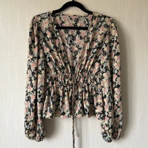 Blommig omlottblus med snörning  - Supersöt blommig omlottblus med v-ringning och långa puffärmar. Blusen har snörning i midjan för en snygg passform och är i mjukt, lätt tyg.