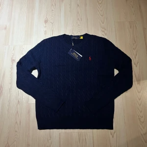 Mörkblå kabelstickad tröja Polo Ralph Lauren - Kabelstickad mörkblå tröja från Polo Ralph Lauren med klassisk rund halsringning och röd broderad logga på bröstet. Tröjan har lång ärm och ribbade muddar för en snygg passform. Perfekt för dig som gillar stilren och tidlös design.